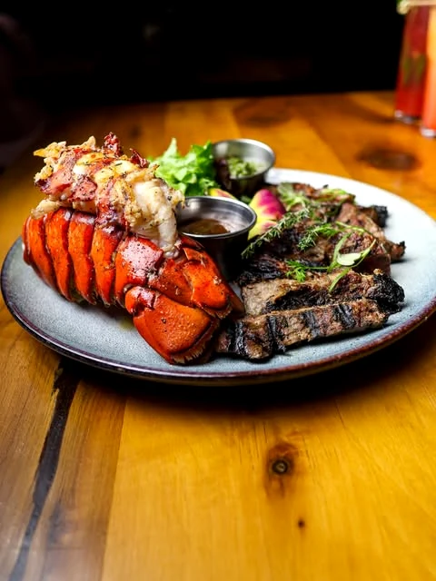Mar y Tierra: New York strip de 12oz con cola de langosta, sobre mesa de madera.